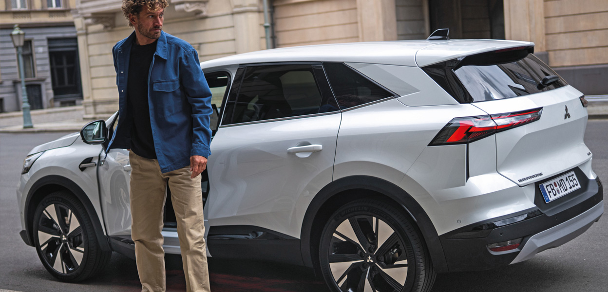 Ein junger Mann, blaues Hemd, beige Hose steigt in einen weißen Mitsubishi Grandes im Hintergrund ein Sandsteingebäude