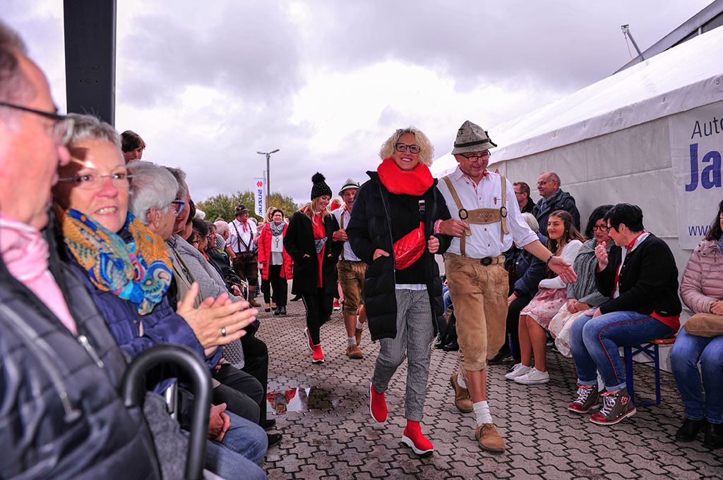 Modenschau im Freien, mit vielen Besuchern, ein Model mit blonden Haaren, rotem Schal, schwarze Jacke, Jeans sowie einem Herrn aus der bayerischen Kapelle Armin Arm über den Laufsteg