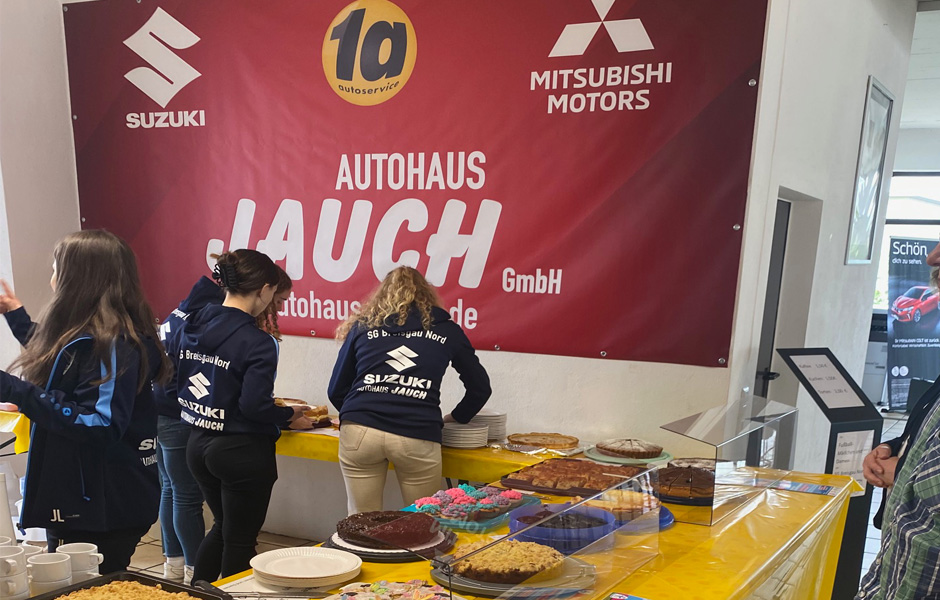 Im Hintergrund eine Plane in rot in der oberen Reihe, das Logo der Firma Suzuki das Logo 1A sowie das Logo der Firma Mitsubishi im unteren Bereich sehen wir vier Jugendliche in Sweatshirts der Firma Autohaus Jauch Sie arbeiten an einer Kuchentheke